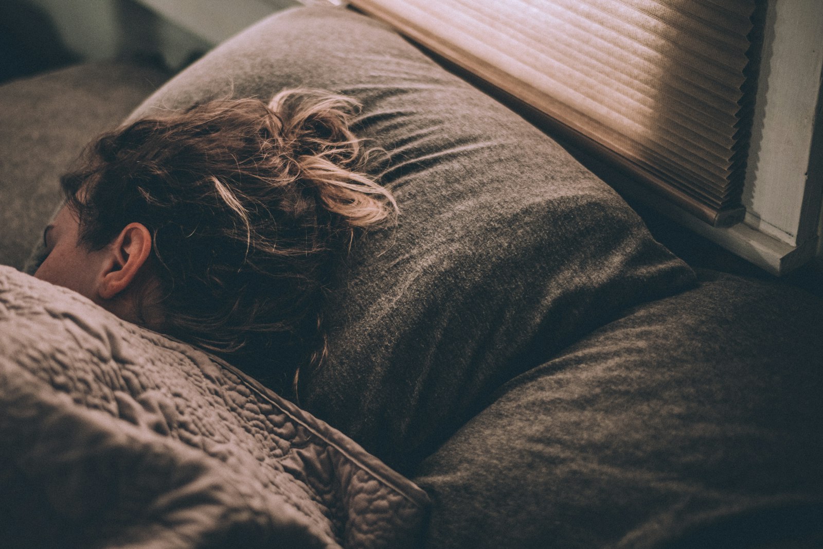 ブラインド越しに日差しが差し込む部屋で枕に顔をうずめて昼寝をする人
